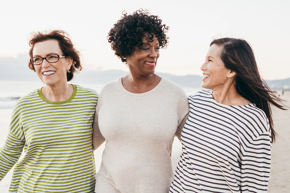 Três mulheres sorriem e caminham juntas na praia.